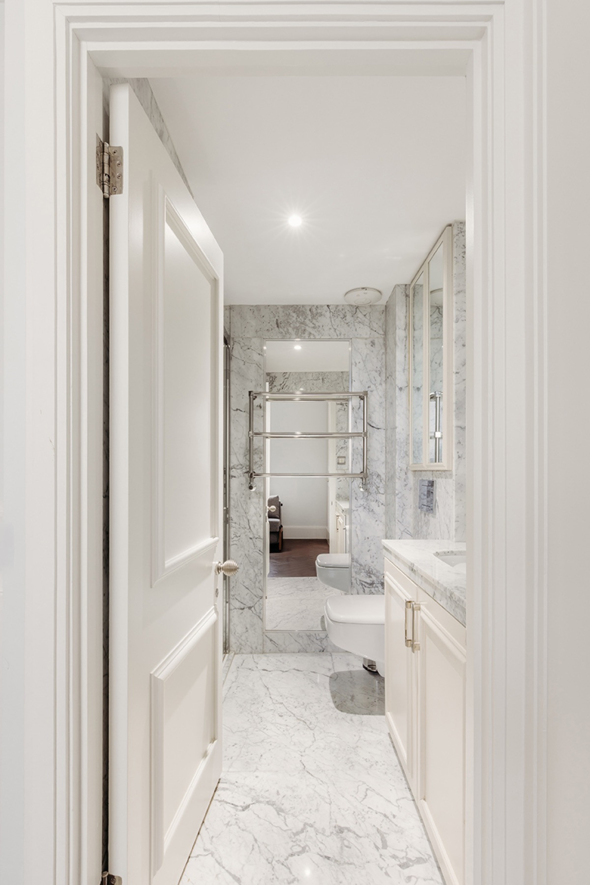 Looking through an open white panelled door into a bathroom lined in white marble, with a wall hung WC, a heated towel rail and a painted vanity unit with a marble top.