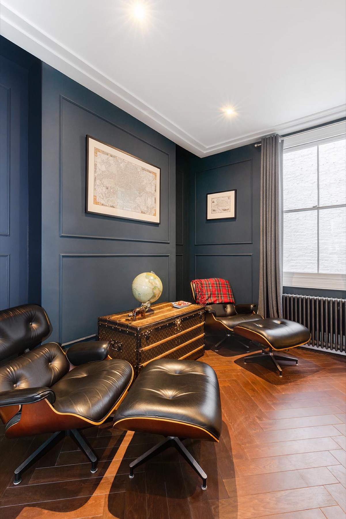 The library at Margaretta Terrace, Chelsea: deep blue walls with panel mouldings painted to match, a white cornice and ceiling, a sash window, herringbone flooring and leather lounge chairs.