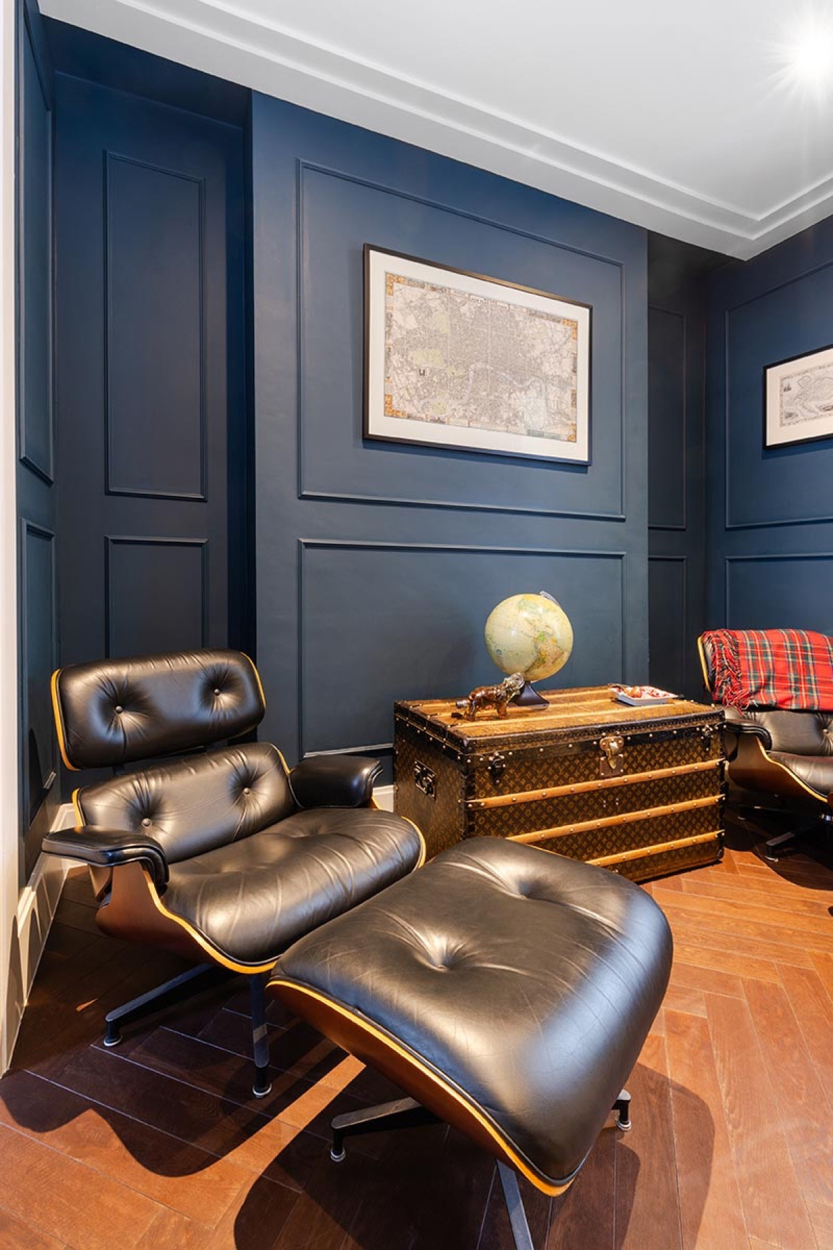 A library with deep blue panelled walls, a white cornice and a herringbone floor, with a black leather lounge chair and footstool, a vintage trunk carrying a globe, and framed maps.
