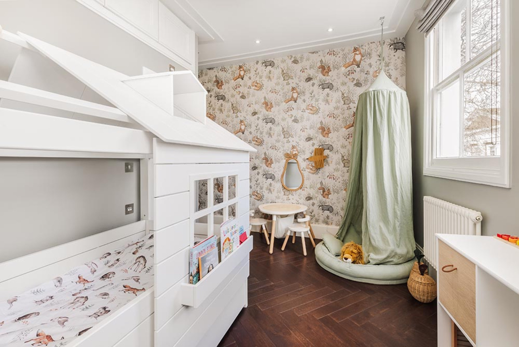 A child's bedroom with a white house shaped bunk bed, woodland animal wallpaper, a sage green play canopy, a dark herringbone floor, a white column radiator and a tall sash window under a moulded cornice.