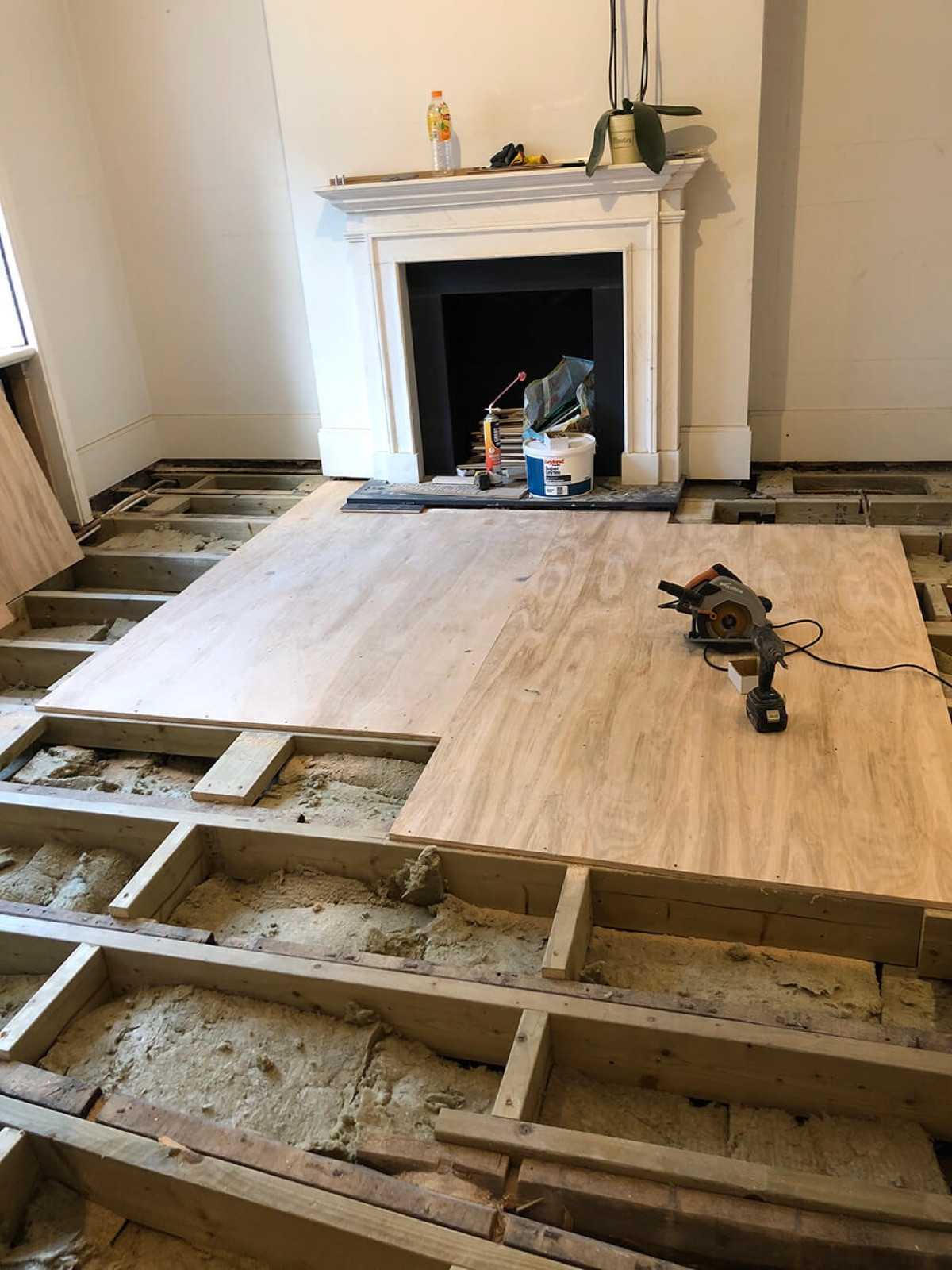 A room in a Victorian townhouse at Margaretta Terrace, Chelsea, during the build: the floor opened up to its joists, insulation laid between them and new plywood boards going down in front of a white fireplace.