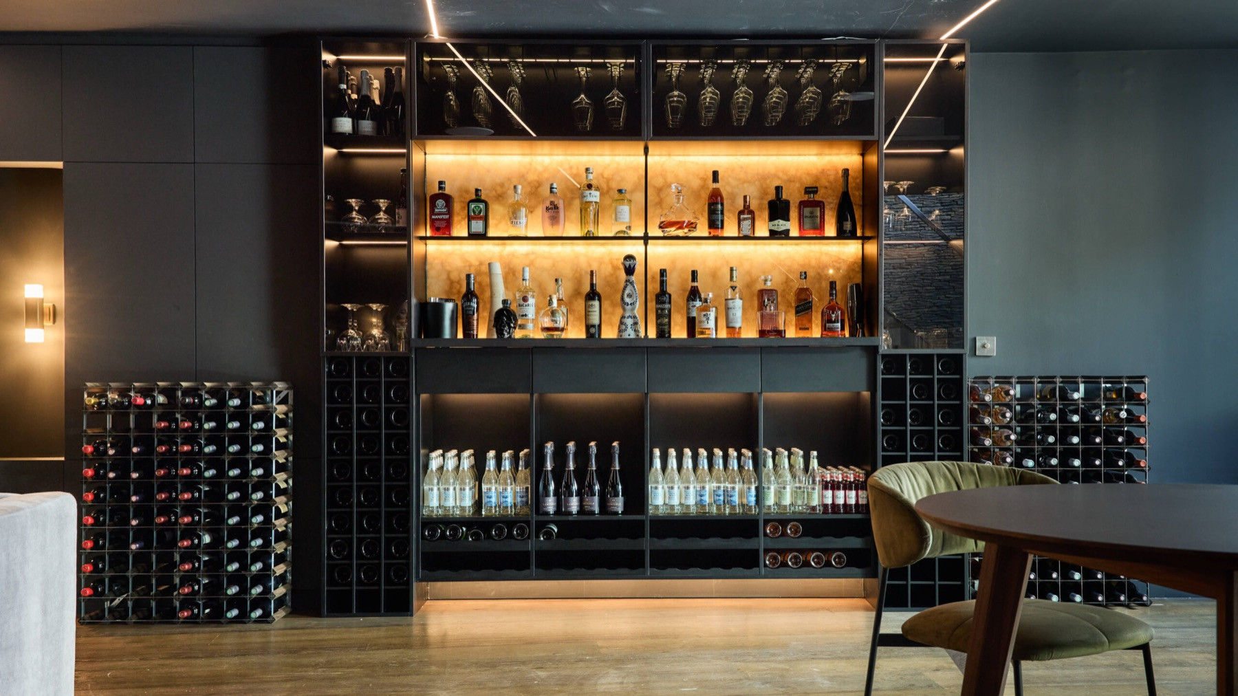 The bar at Ellerby Street, Fulham: dark joinery and wine racks around shelves lit warmly from behind, with a green velvet chair at a round table.