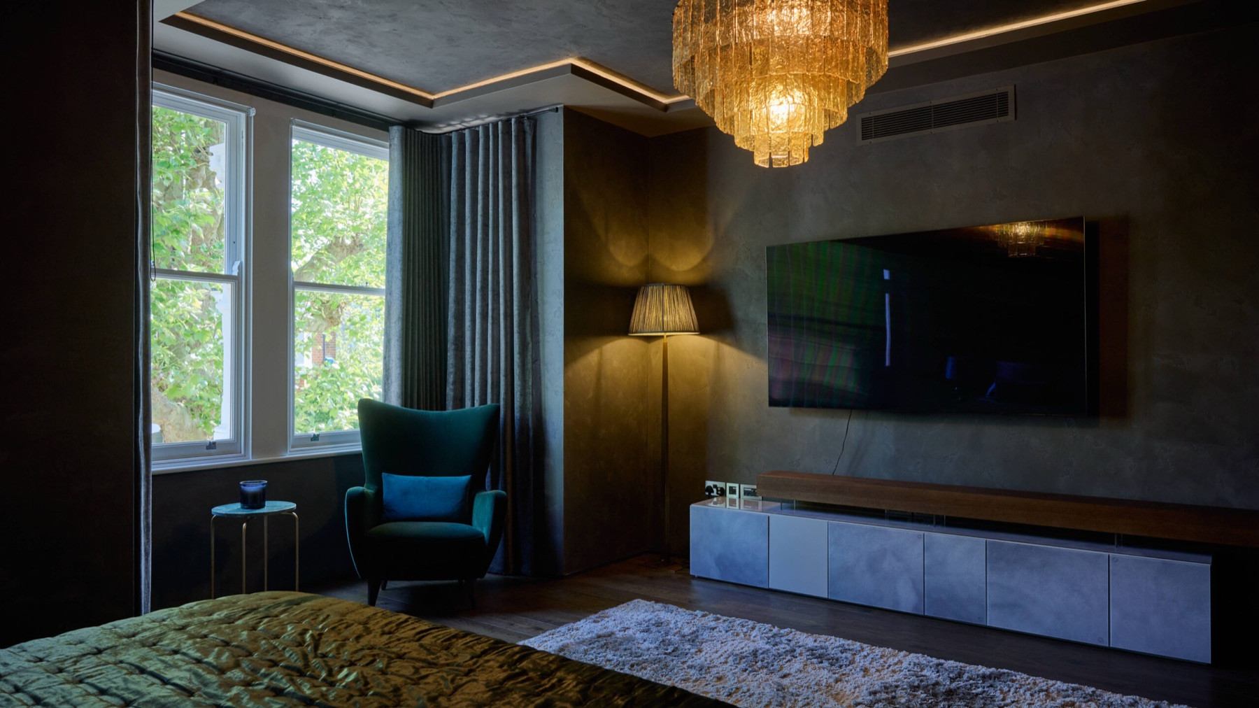 A bedroom at Ellerby Street, Fulham, lit in layers: a tiered amber glass chandelier, concealed light around the ceiling and a floor lamp beside the window.