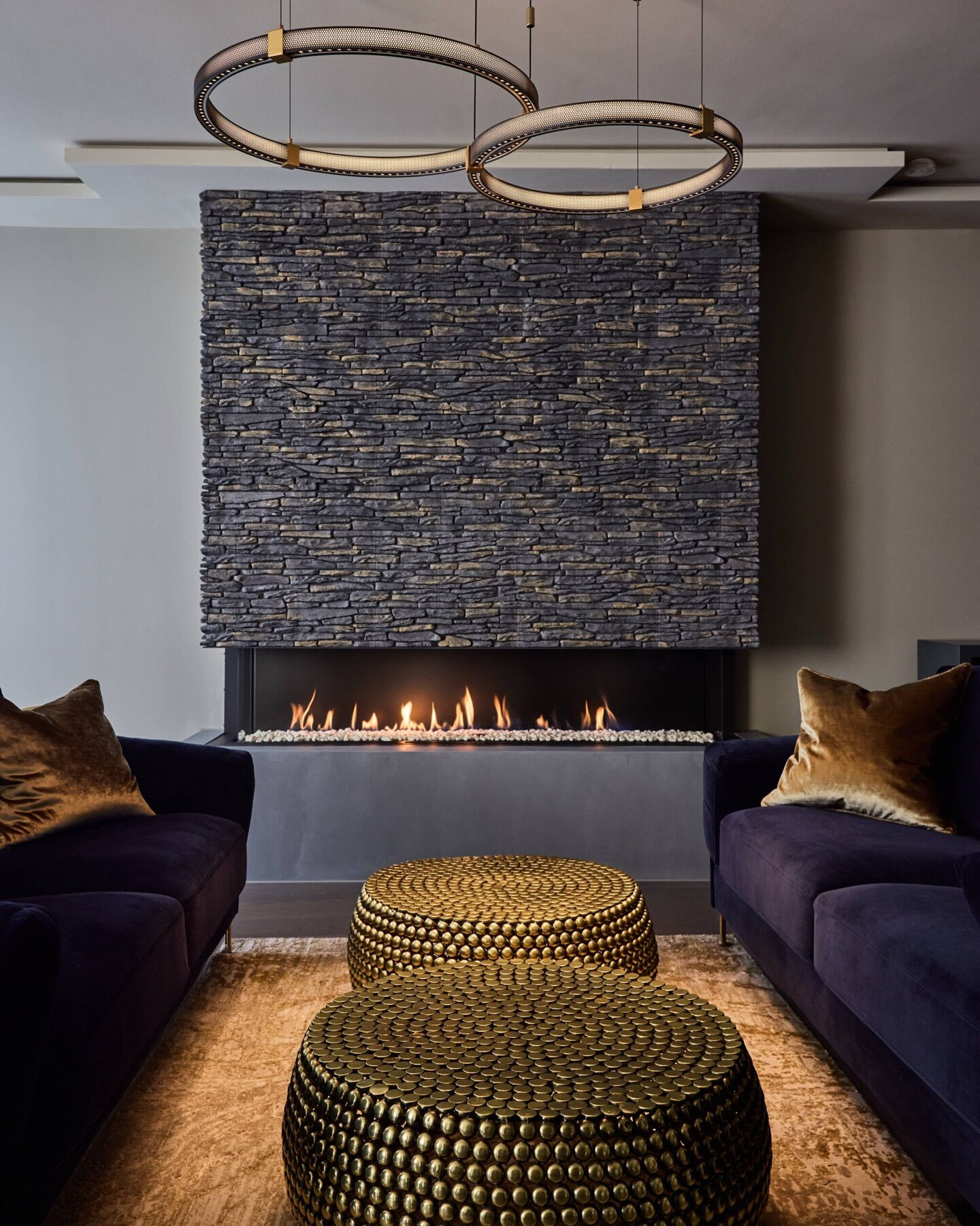 A dark living room with a long, low fire set into a wall clad in stacked stone, two ring pendant lights, navy velvet sofas and two brass studded tables on a rug.