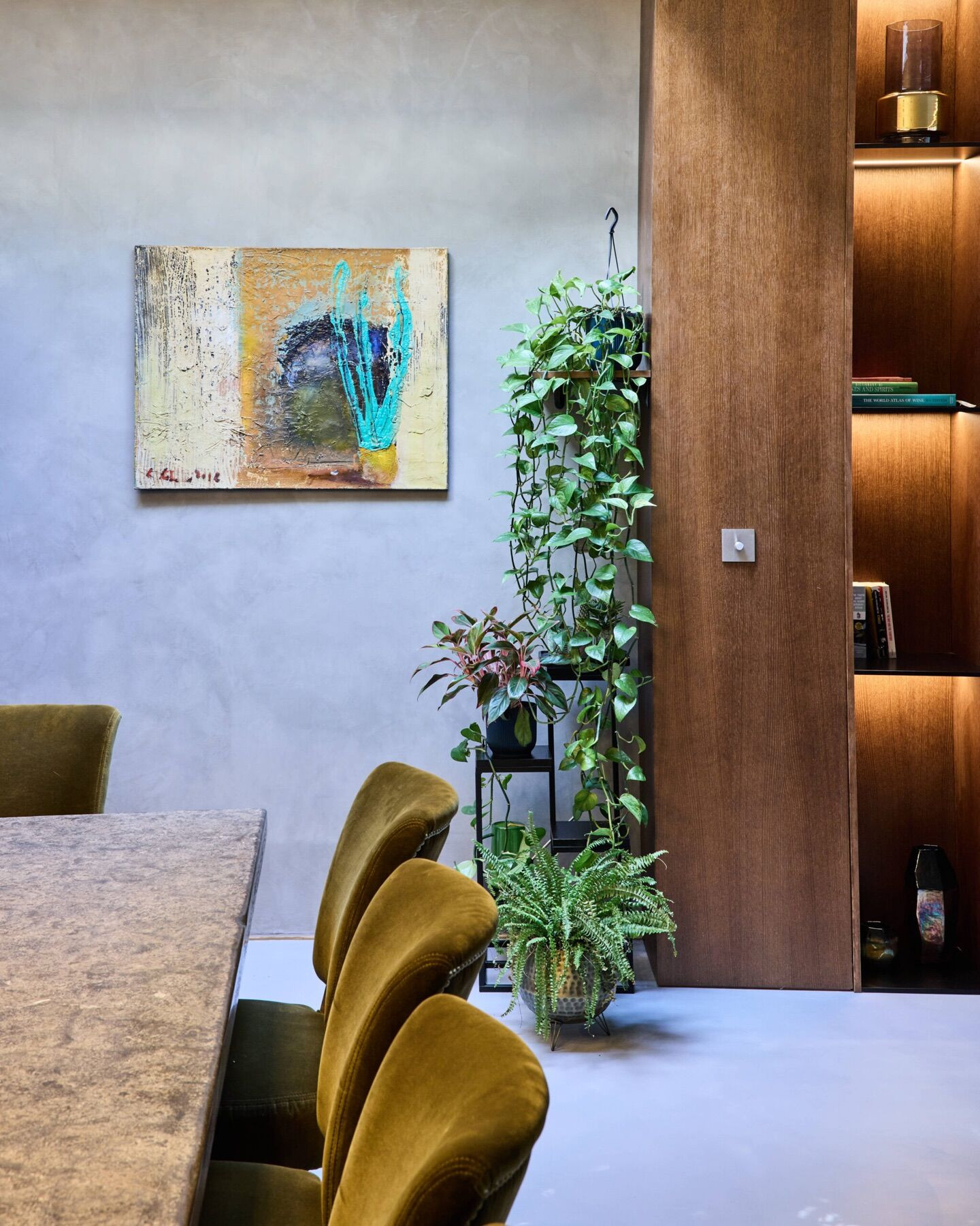 A finished dining area with mottled grey plaster walls, an abstract painting, plants on a stand, olive velvet chairs at a stone topped table, and dark wood joinery with lit shelves and a metal light switch.