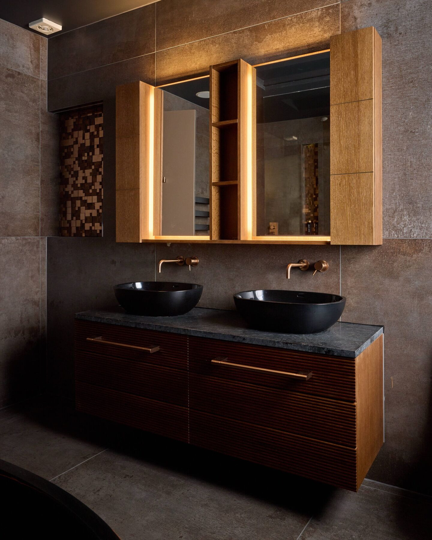 A wall hung double vanity with fluted timber drawers and long copper coloured handles, a dark stone top with two black vessel basins and wall mounted copper coloured taps, under a lit oak mirror cabinet, with large dark tiles and a mosaic niche