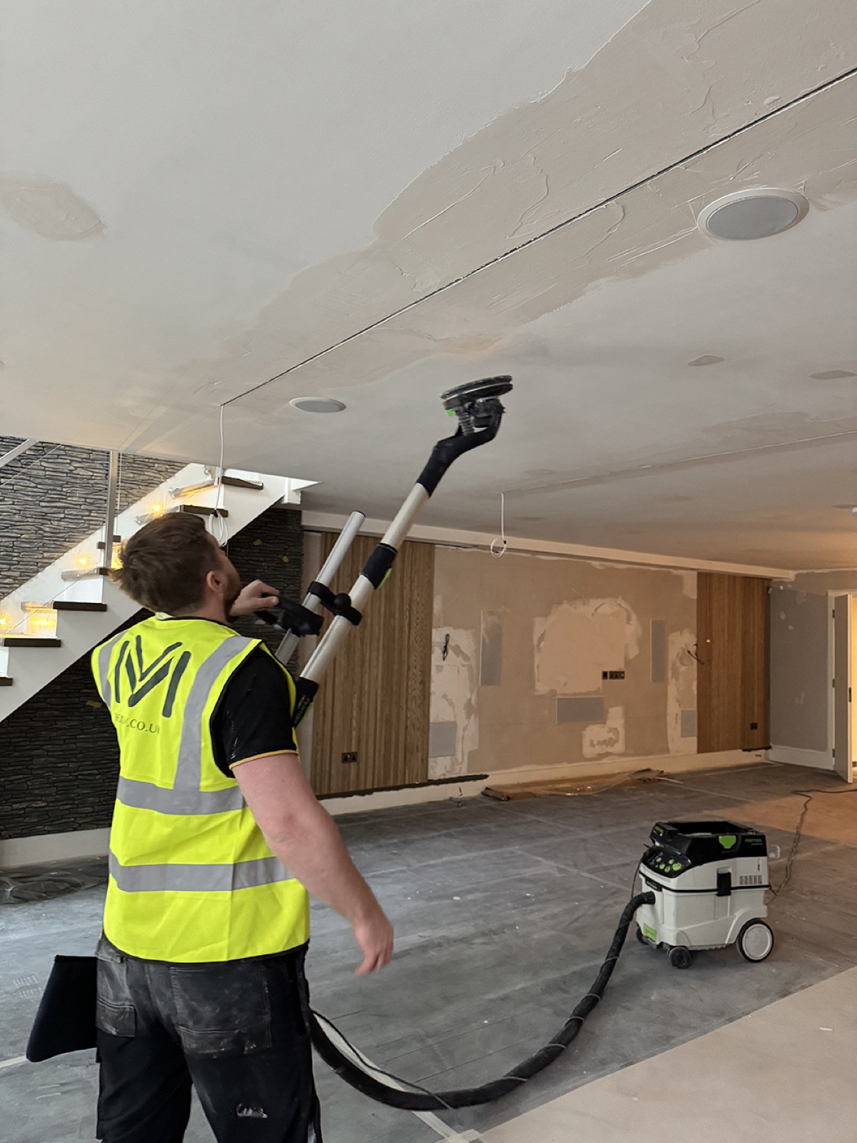 A site operative in a high-visibility vest sanding a ceiling with a long-handled sander connected by a hose to a dust extractor, in a large open room with the floor covered in protection and a staircase behind.