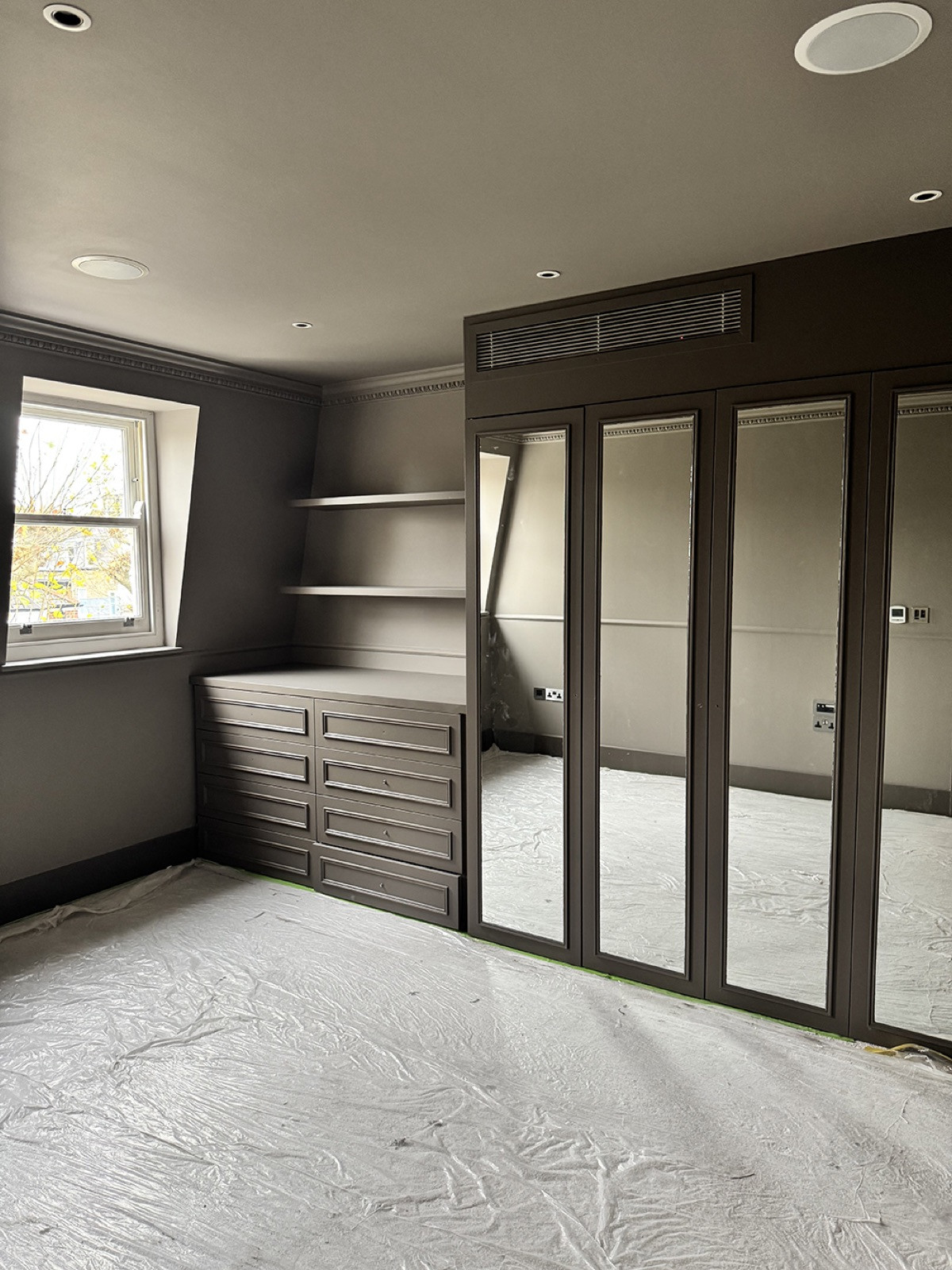 A room in the final stage of work: fitted wardrobes with mirrored doors, a chest of drawers and shelves painted a dark taupe, a sash window, and the floor still under protective sheeting.