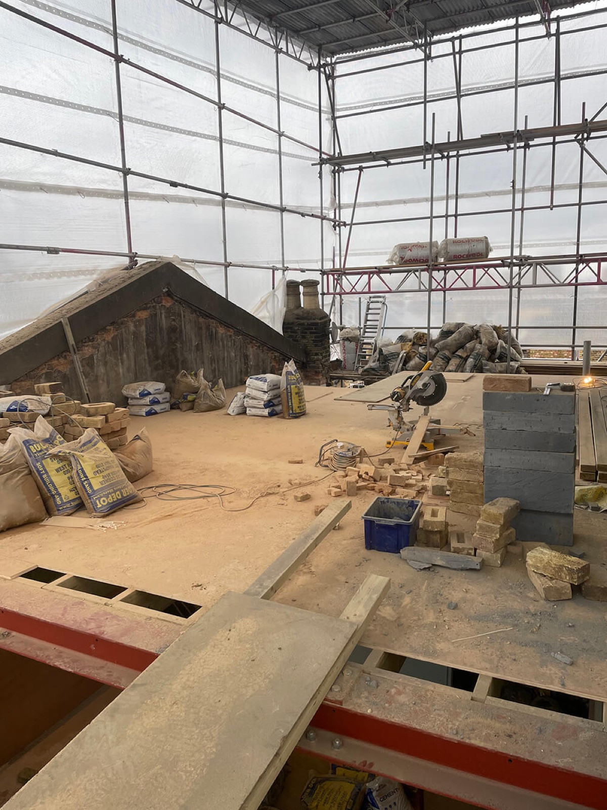 A new floor deck on red steel beams at roof level, under scaffolding wrapped in white sheeting, with bricks, sand bags, a mitre saw and the old roof's gable and chimney pots beside it.