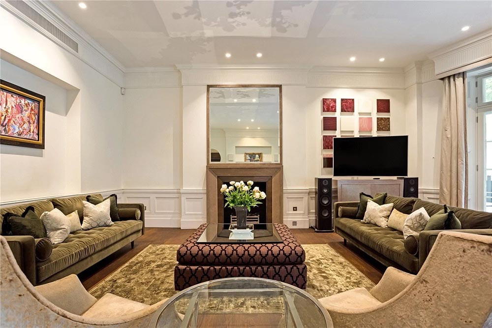 A living room with panelled walls, a deep cornice, a stone fireplace under a tall mirror, a dark wood floor, two olive velvet sofas and a patterned ottoman.