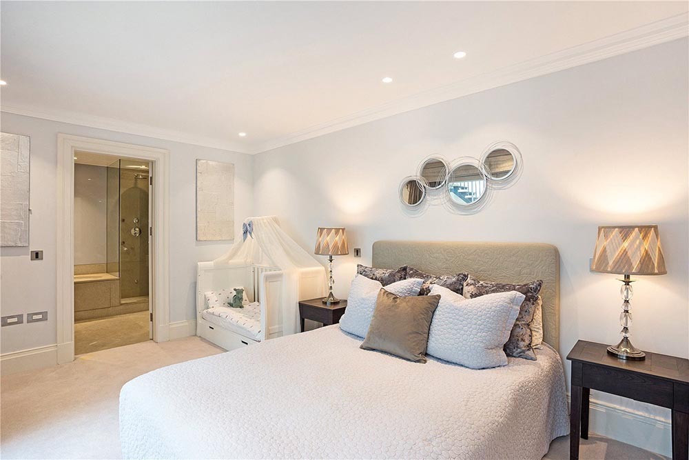 A pale carpeted bedroom with a cornice and downlights, an upholstered bed between two bedside lamps, and an en suite with a glass shower seen through the open door.