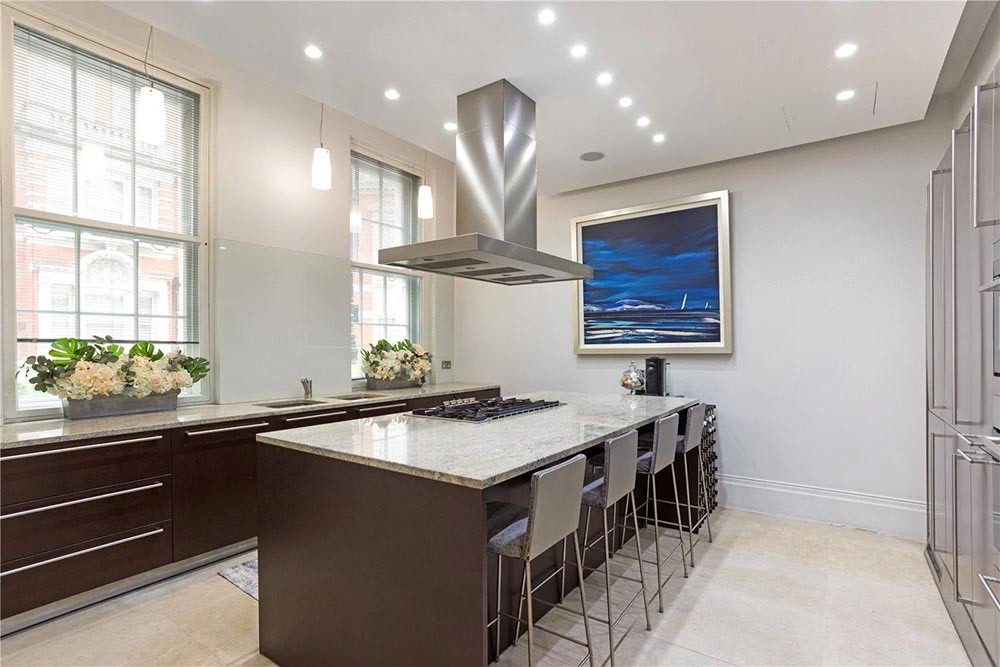 A kitchen with dark wood units and long steel handles, pale speckled stone worktops and an island with a gas hob under a steel chimney hood and four bar stools, beside tall sash windows, on a pale stone floor