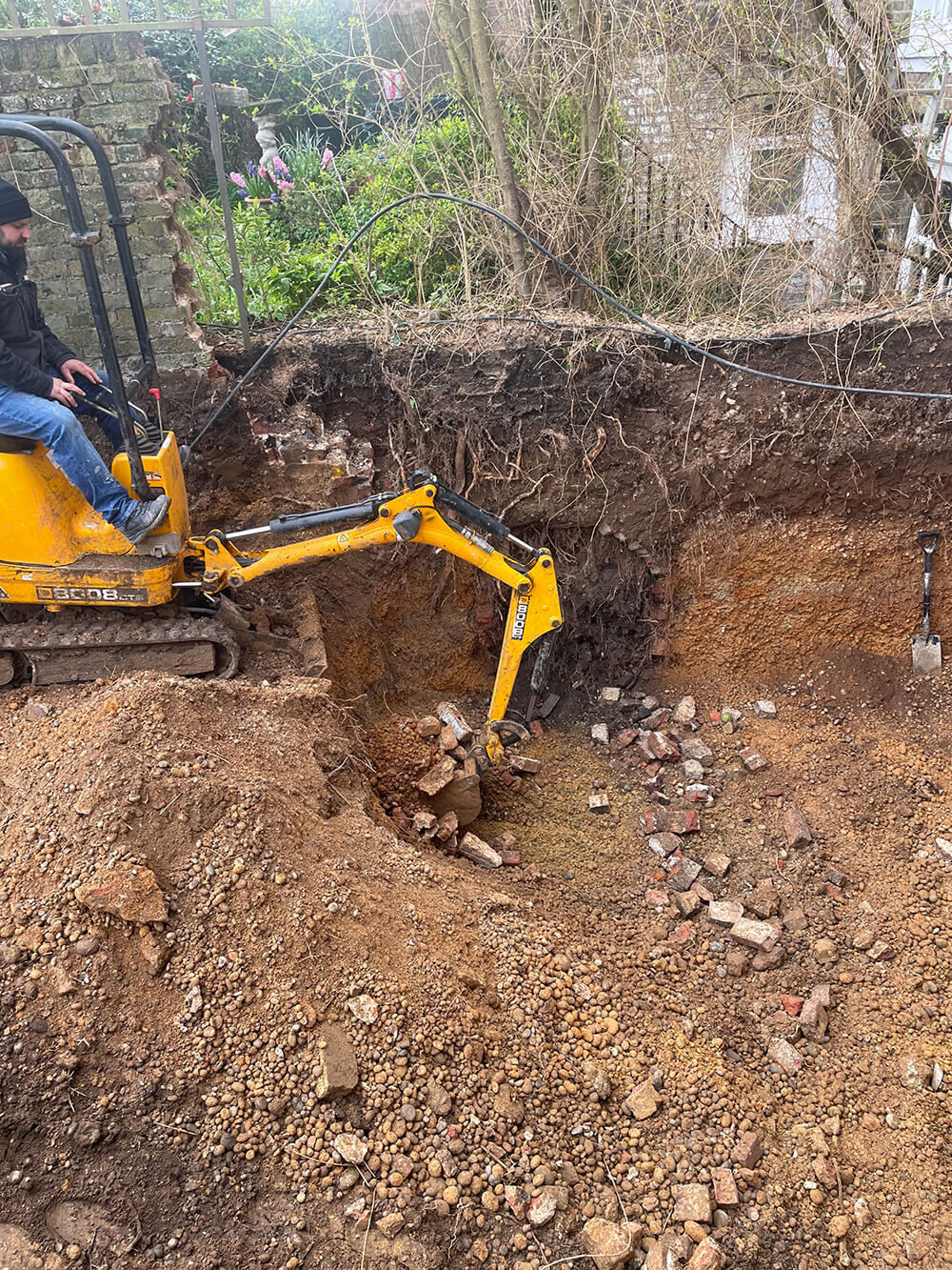 A mini excavator and its operator digging out a deep hole for a rear basement extension at St Germans Place, Blackheath, with old brickwork exposed in the side of the dig and a garden wall and planting just above it.
