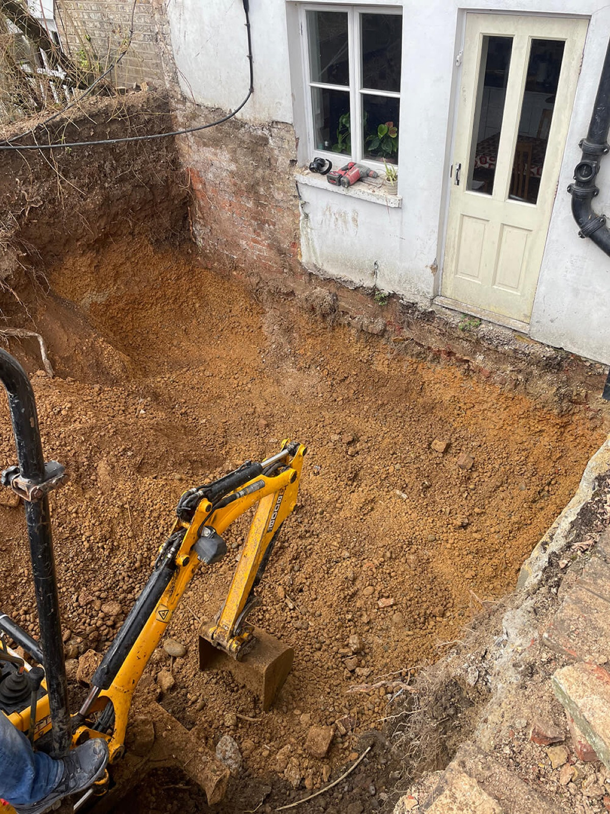 A mini digger excavating the garden behind a house for a rear extension, the ground dug out below a window and a glazed back door, exposing the brickwork beneath the render.