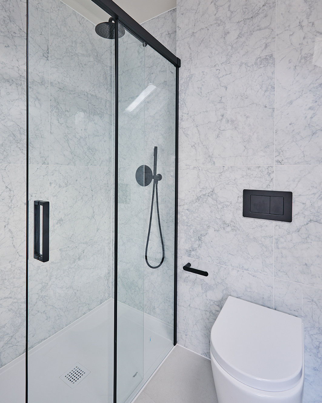 A walk in shower with a black framed sliding glass door, a black rain head and round concealed valve on white tiles veined like marble, beside a wall hung toilet with a black flush plate on the wall above it